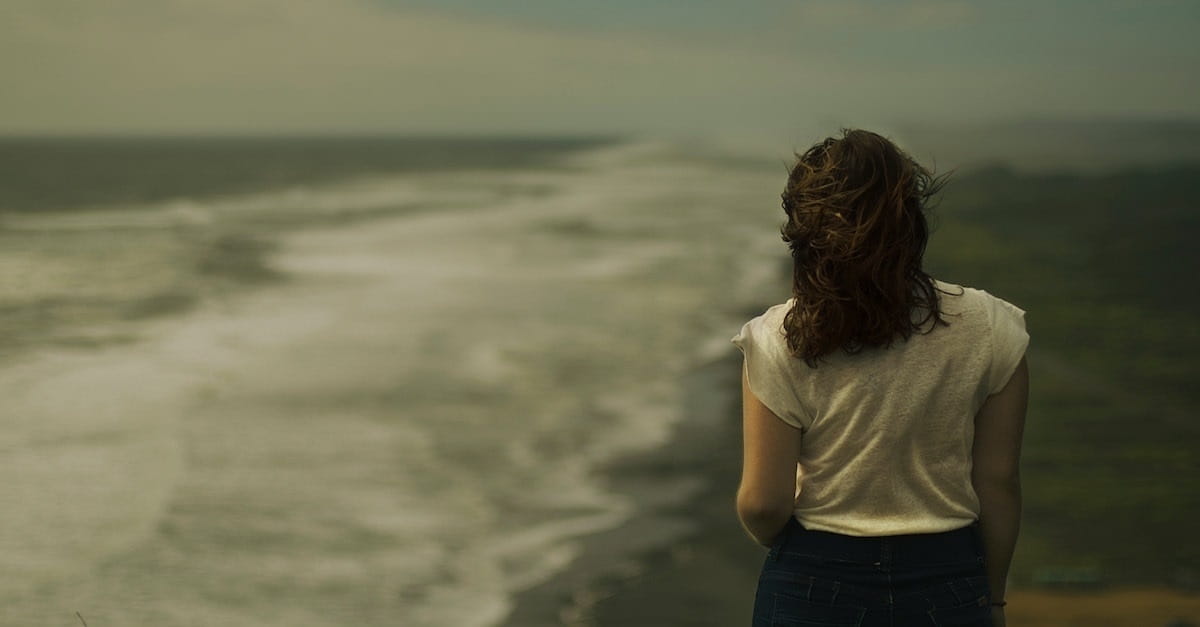 A woman walks away from the camera down a beach in thought under grey skies