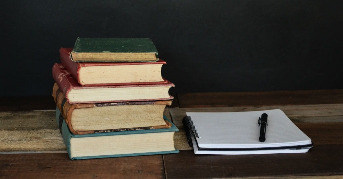 Five books stacked on table next to notebook with paper