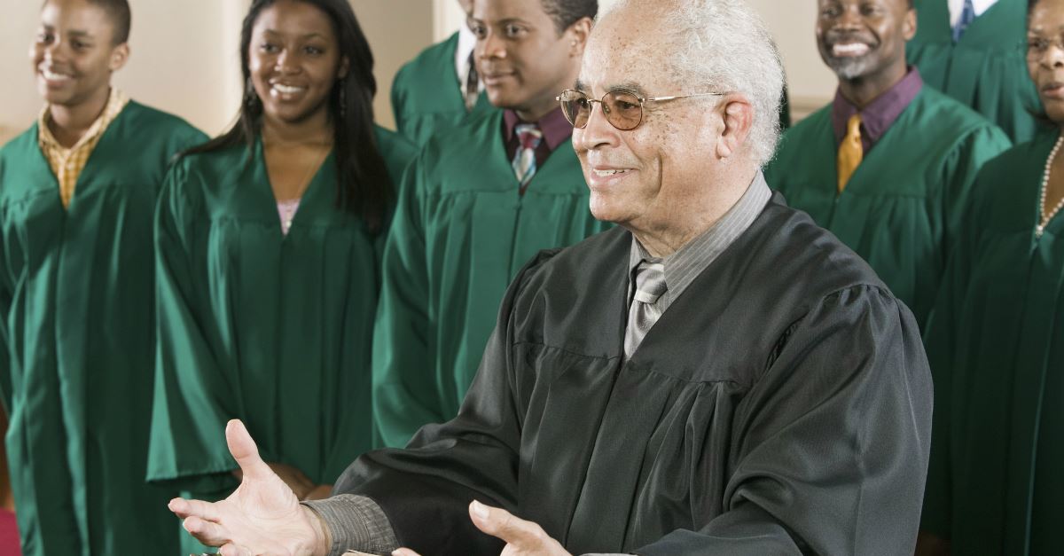 older pastor with choir in background, smiling, hands open, preaching