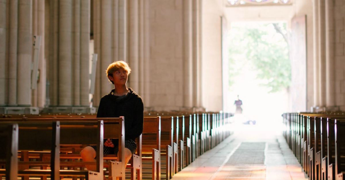 young man sitting alone in empty church sanctuary looking up, shadow of person walking through sunlit doorway behind him