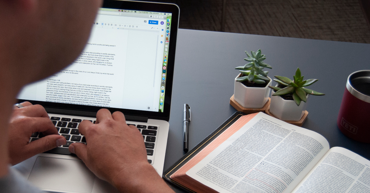 over shoulder man reading bible while typing newsletter at laptop work pastor