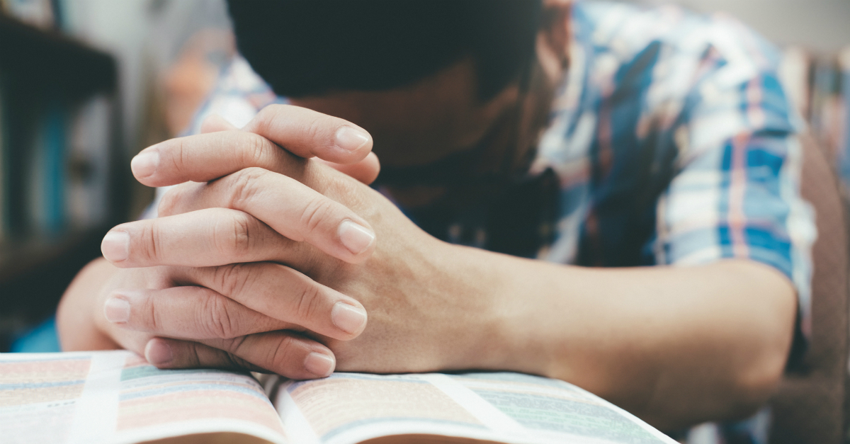 man with head down hands folded on open Bible praying