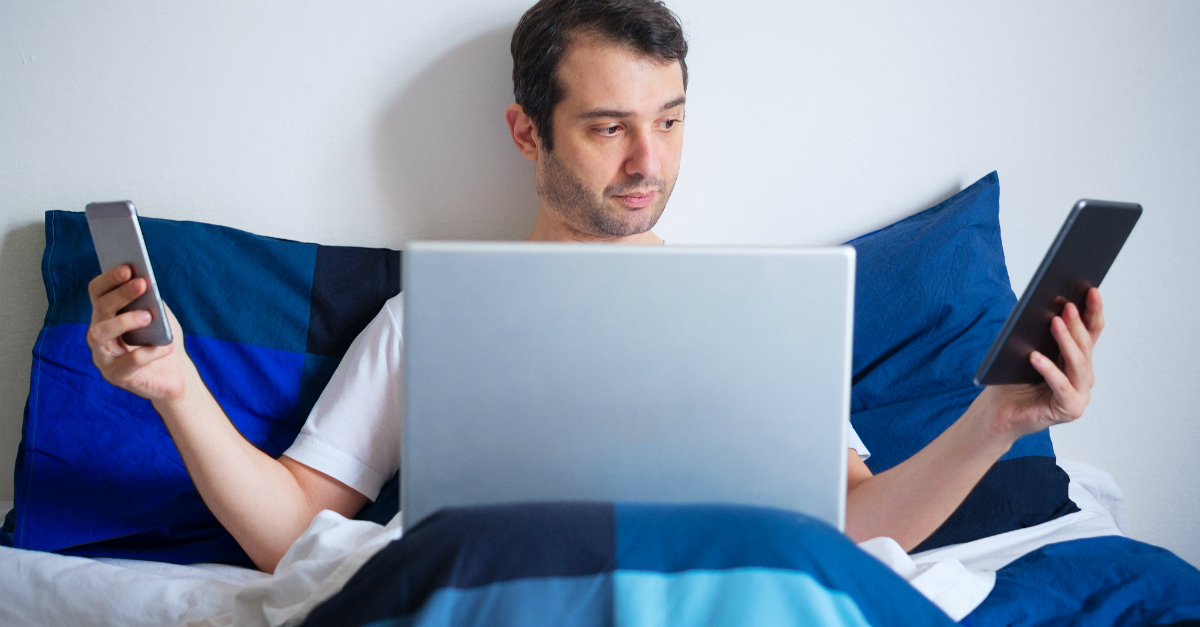 man tired in bed on two phones and laptop