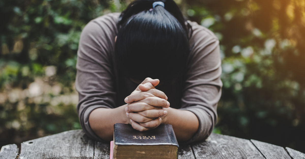 woman with head down on bible on table praying solemnly