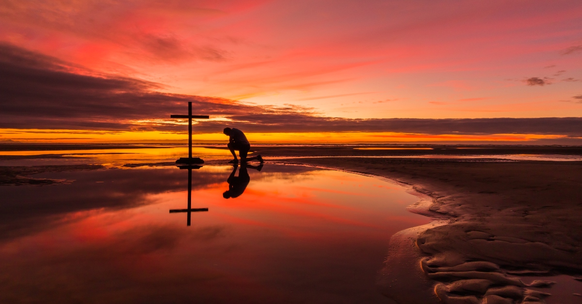 man kneeling before cross on beautiful beach sunset, every knee shall bow meaning