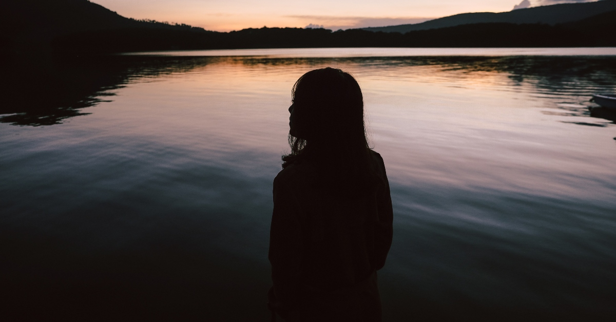 woman standing silently before a lake as sun sets