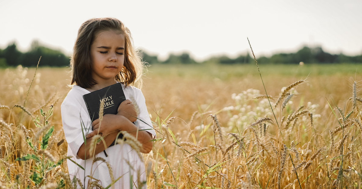 little girl holding Bible outside in a field with her eyes closed