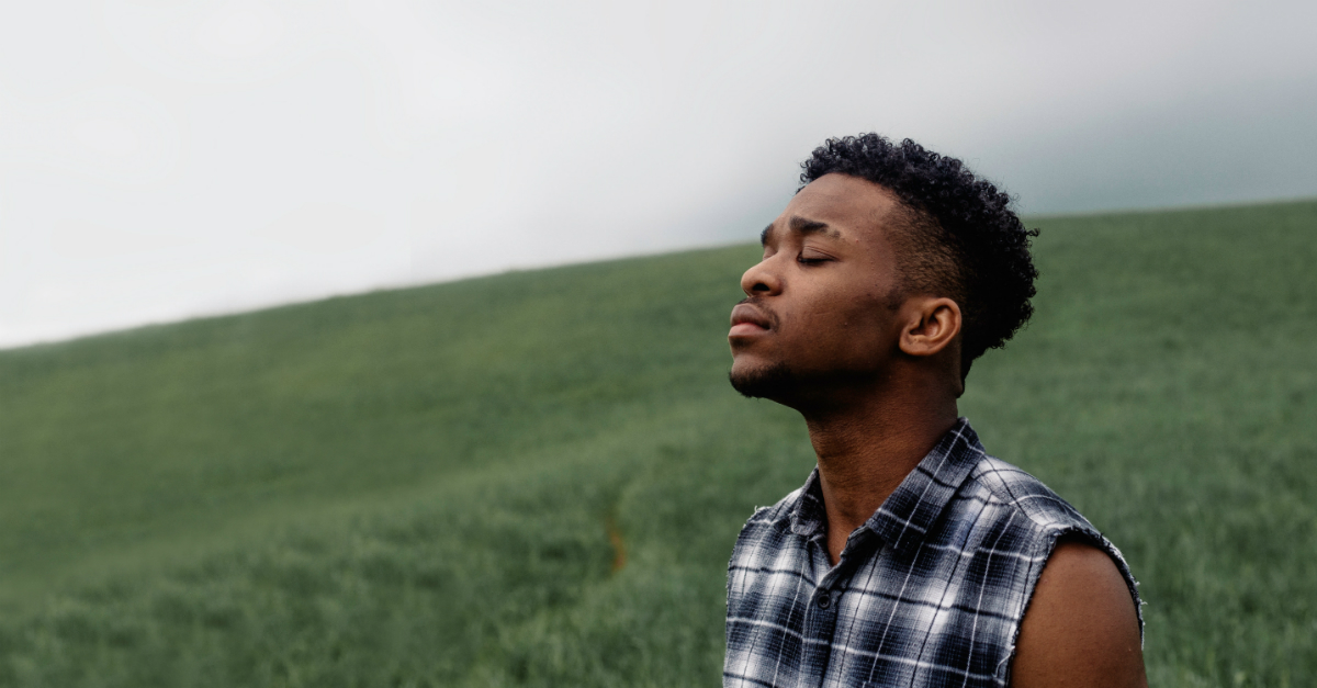 man standing on grassy hill with eyes closed in prayer