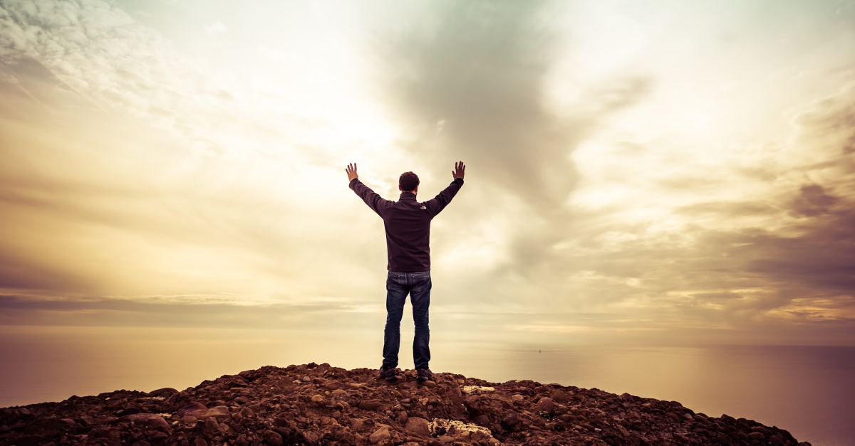 man on hilltop arms raised at sunset in praise