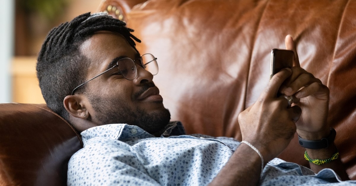 relaxed biracial man lying on leather couch using mobile phone