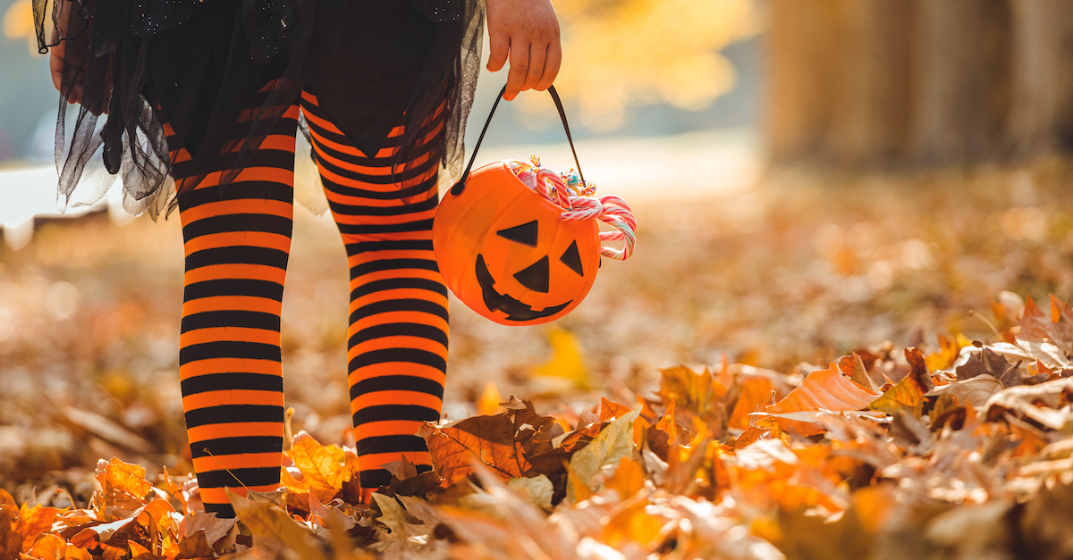 little girl trick-or-treating walking down sidewalk with leaves and holding pumpkin bag with candy