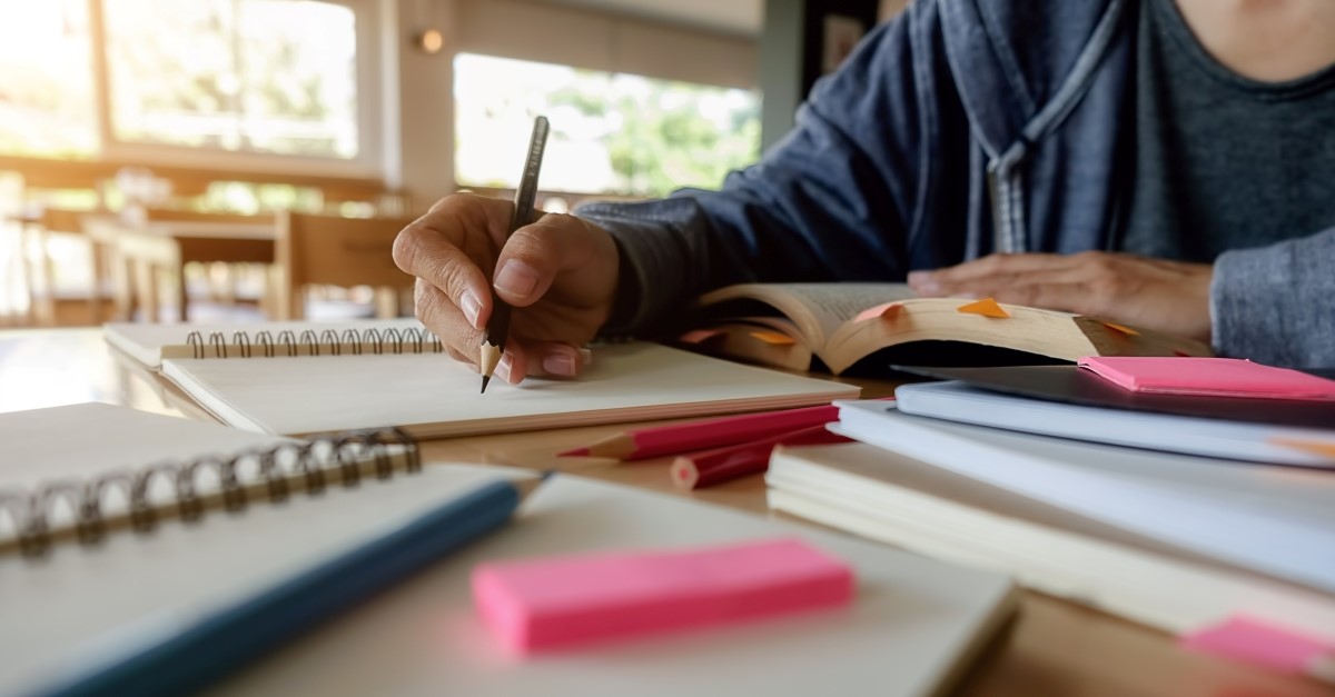 man writing with lots of textbooks and notepads around him